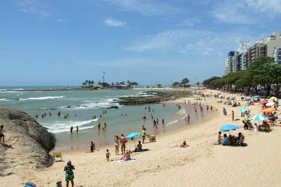Uma praia cheia de gente se banhando e tomando sol, com guarda-sóis e edifícios ao fundo.