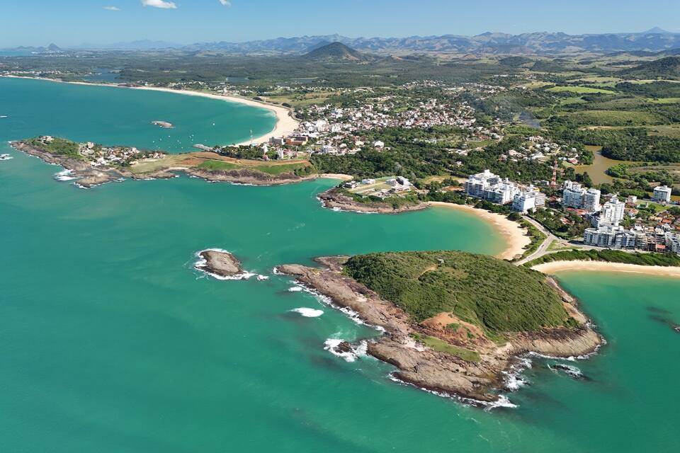 Vista aérea de uma vila costeira com praia, ilhas rochosas e montanhas ao fundo.