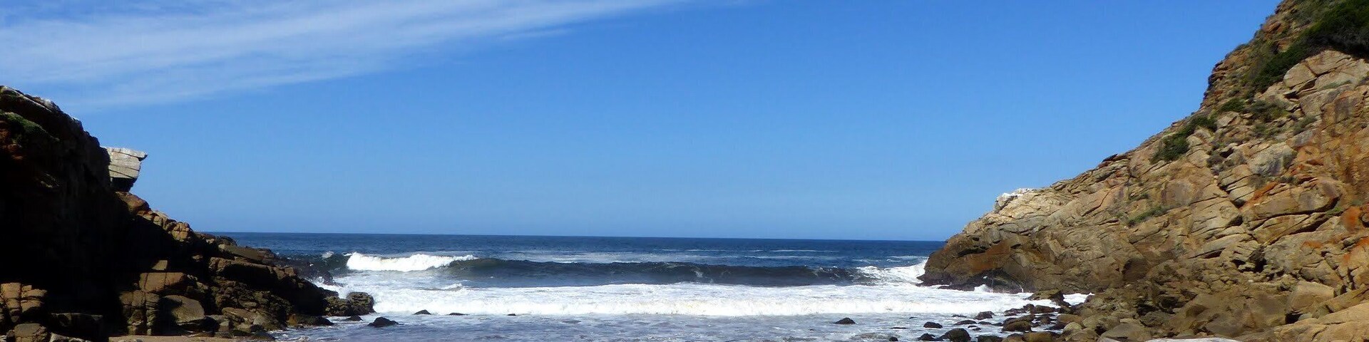 Uma praia rochosa com ondas quebrando na costa, sob um céu azul límpido.