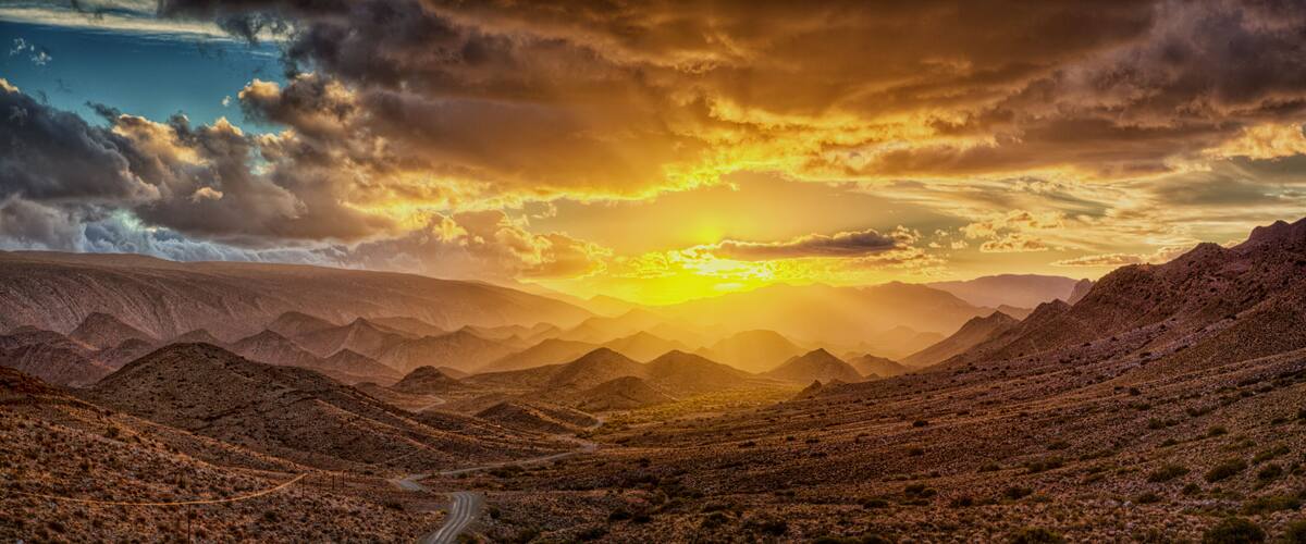 A desert landscape with mountains and a winding road under a dramatic sunset.