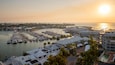 A marina with numerous boats docked, surrounded by buildings and palm trees, during sunset.