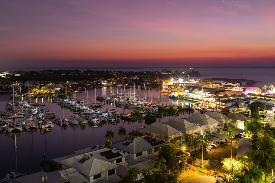 A marina at sunset with boats docked, waterfront buildings, and palm trees.