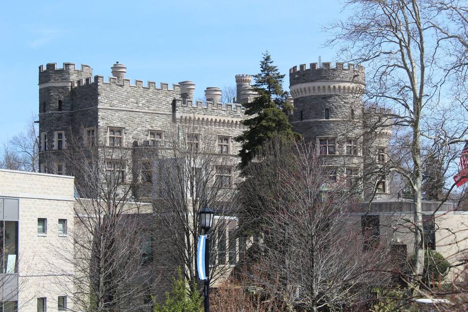 A castle-like building with multiple towers and turrets, surrounded by trees and modern buildings.