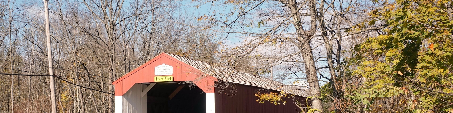 A covered bridge with a red roof and white walls, surrounded by trees and a stone wall.