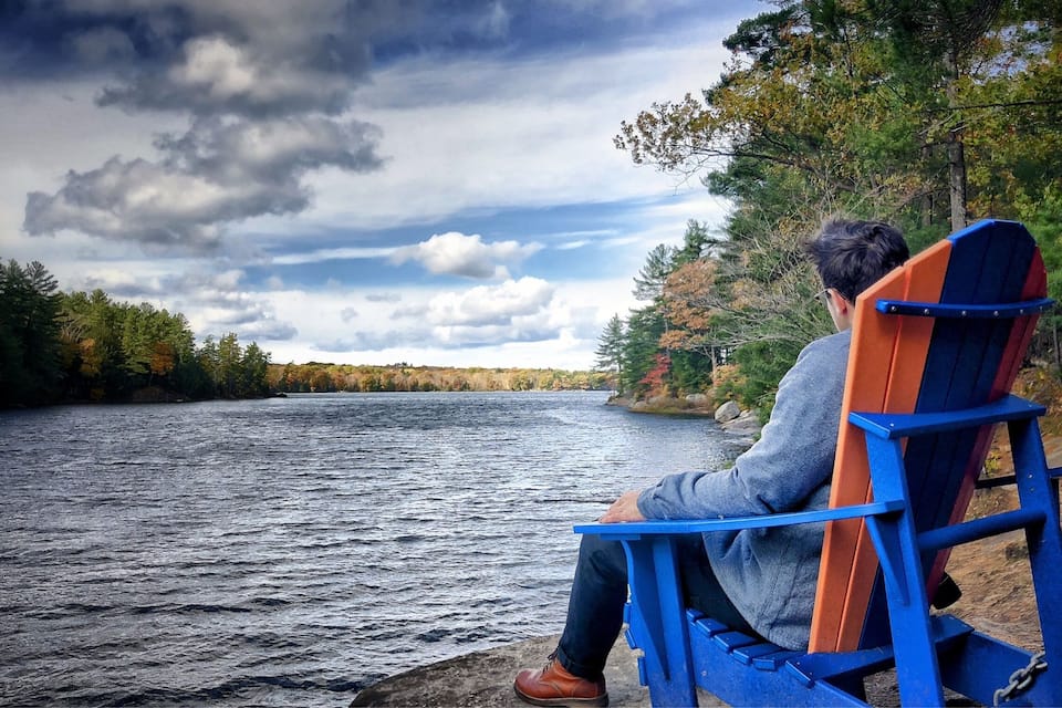 A person sitting on a blue and orange Adirondack chair by a lake with autumn foliage in the background.