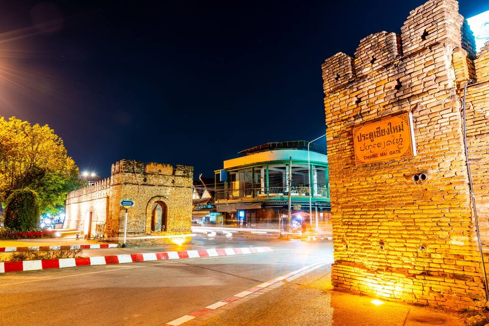 Una porta històrica amb un rètol, un edifici modern i un carrer ple d'arbres de nit.
