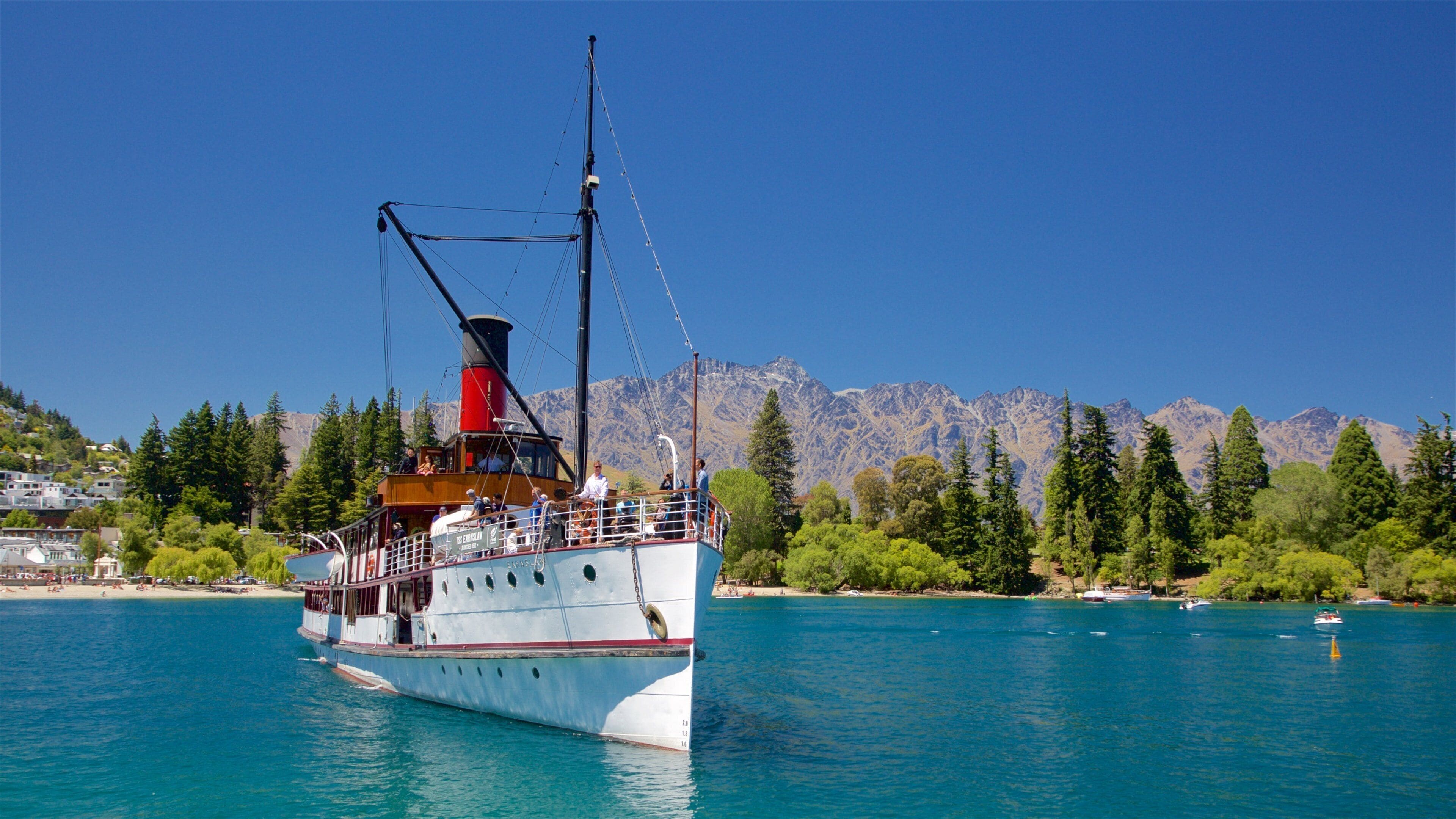 A large white boat with a red funnel and cabin, sailing on a turquoise lake with mountains in the background.