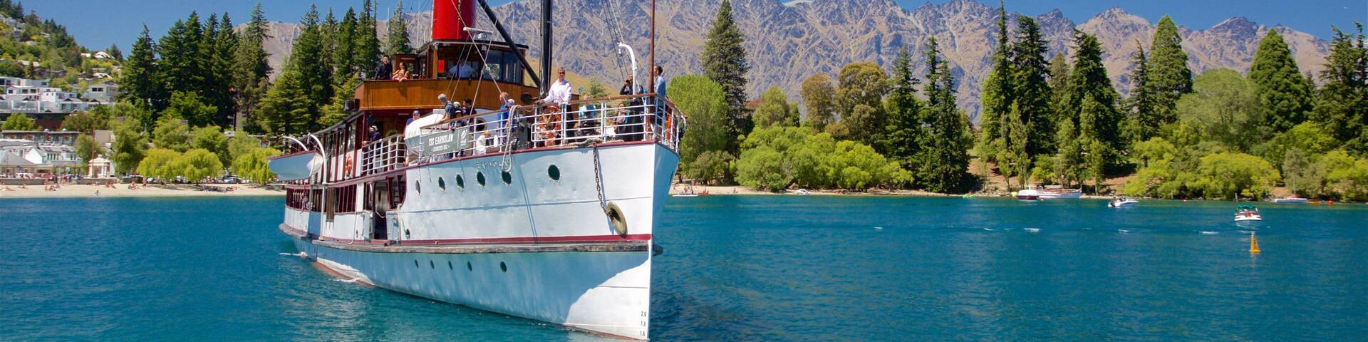 A large white boat with a red funnel and cabin, sailing on a turquoise lake with mountains in the background.