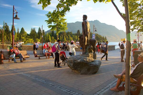 A park with a statue, benches, people, and a lake with mountains in the background.