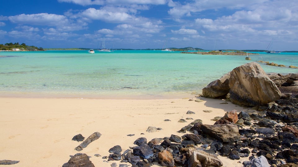 A beach with clear turquoise water, a large rock in the foreground, and sailboats in the distance.