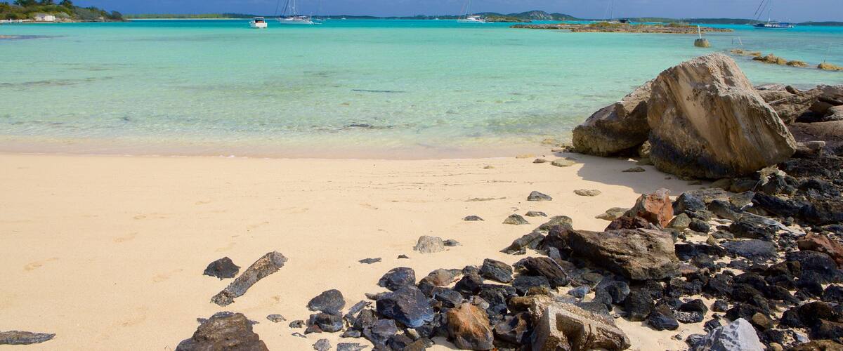 A beach with clear turquoise water, a large rock in the foreground, and sailboats in the distance.