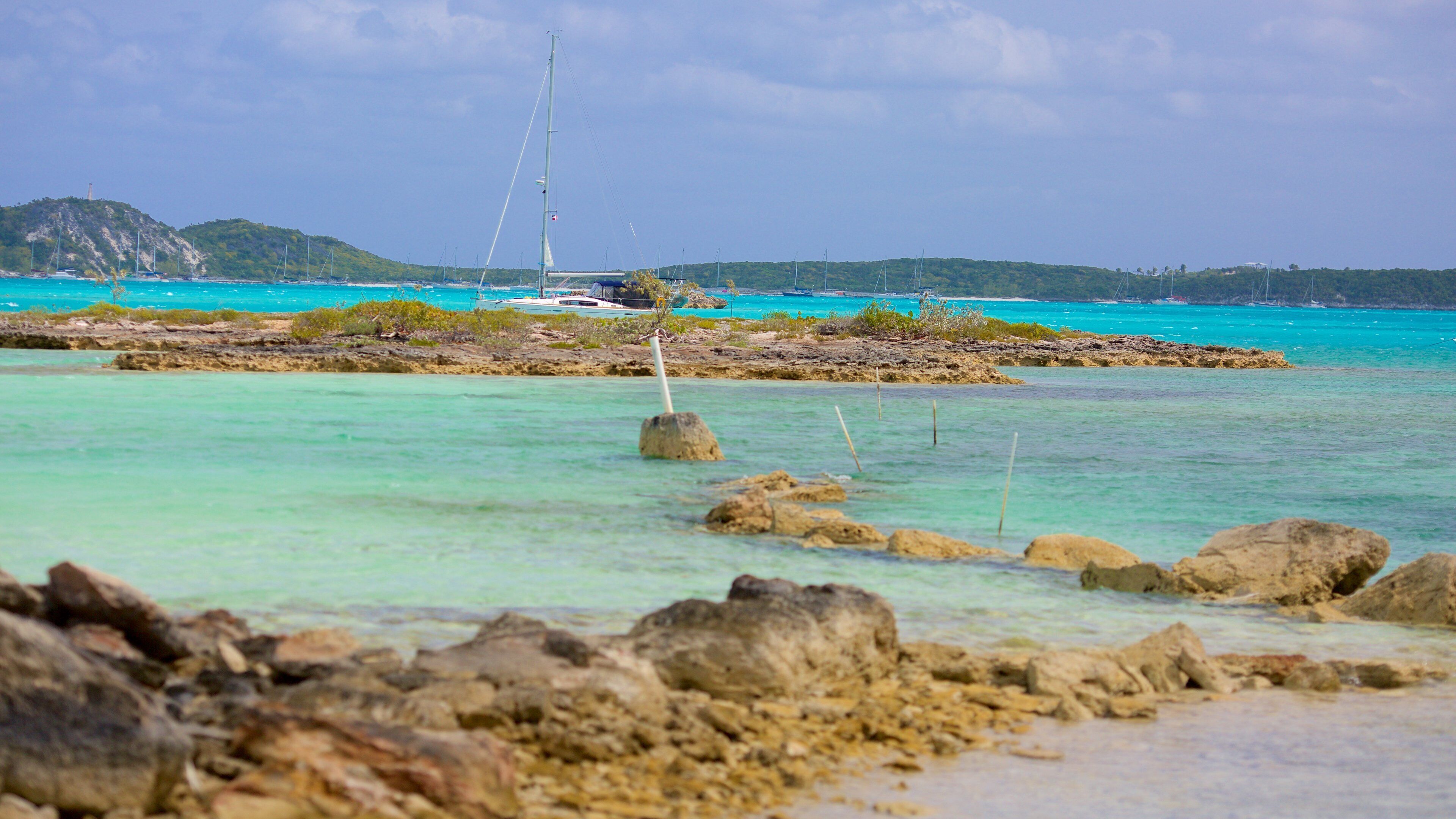 A coastal scene with clear turquoise waters, rocky shoreline, and a sailboat anchored near a small island with a lighthouse.