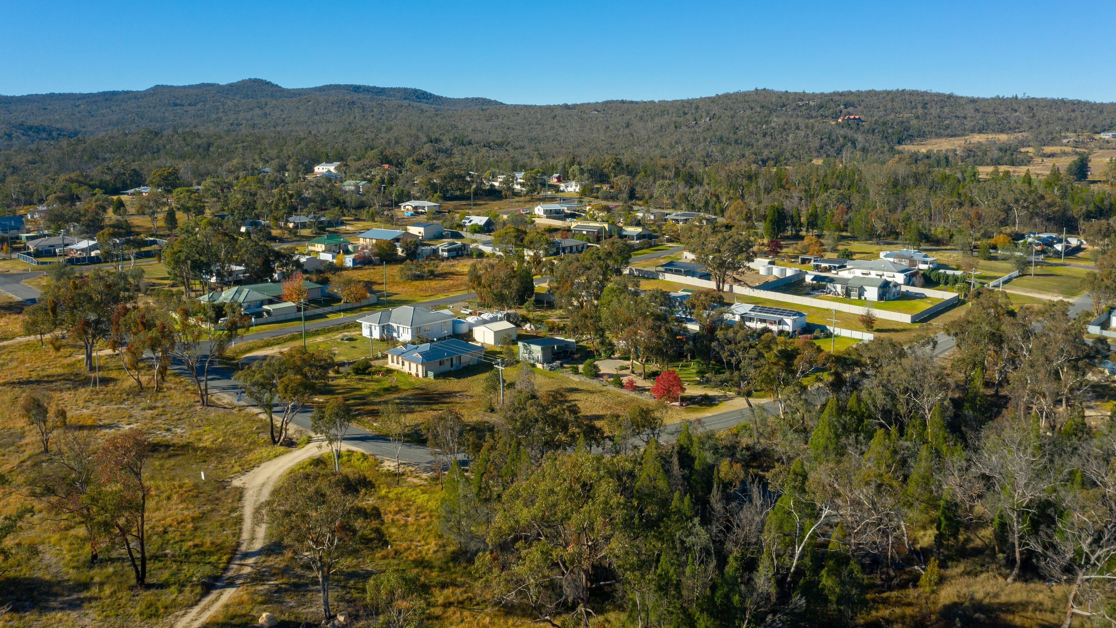 An aerial view of a residential area with houses, roads, and a hilly forested background.