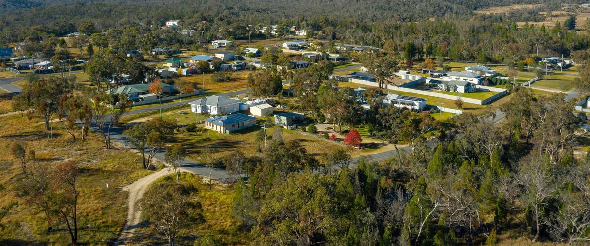 An aerial view of a residential area with houses, roads, and a hilly forested background.