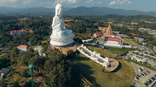 Aerial view of a majestic white Guan Yin statue stands serenely amidst lush greenery and traditional Thai architecture, Guan Yin Statue, Chang Wat Chiang Rai, Thailand.