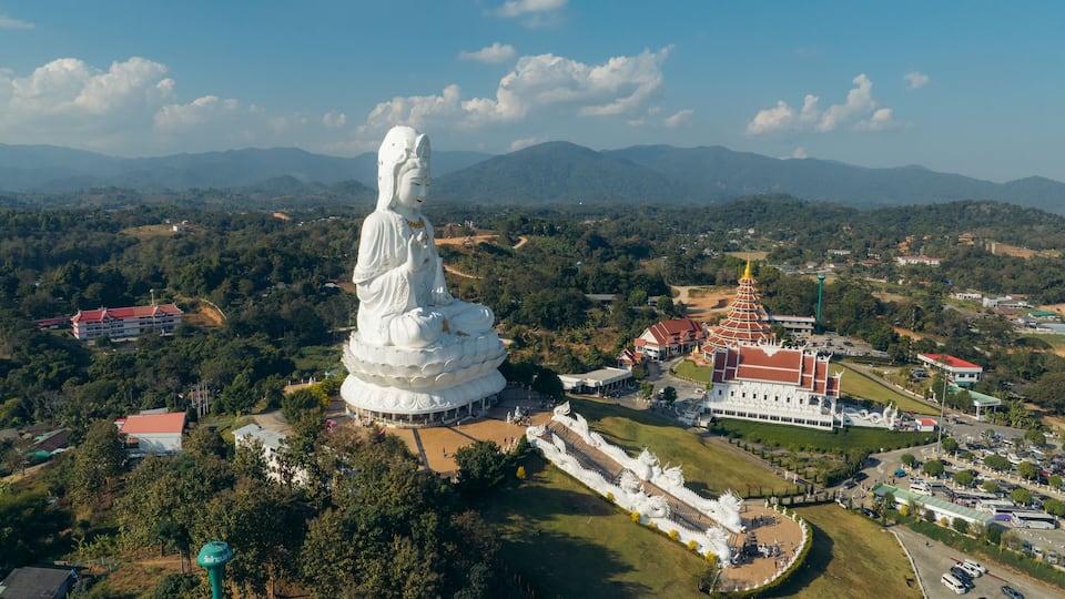 A large white statue of a seated figure surrounded by a temple complex and lush greenery.