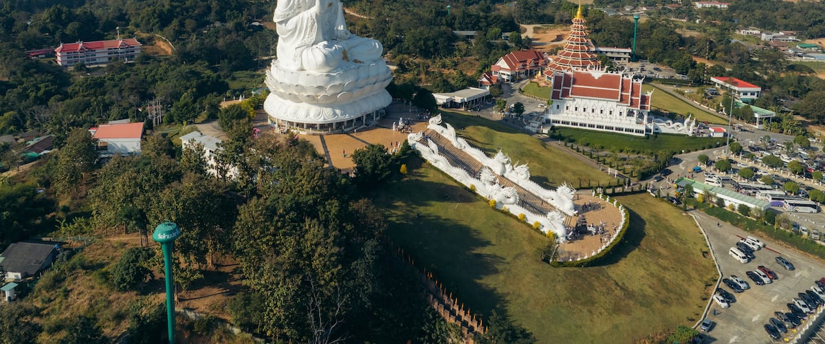 A large white statue of a seated figure surrounded by a temple complex and lush greenery.
