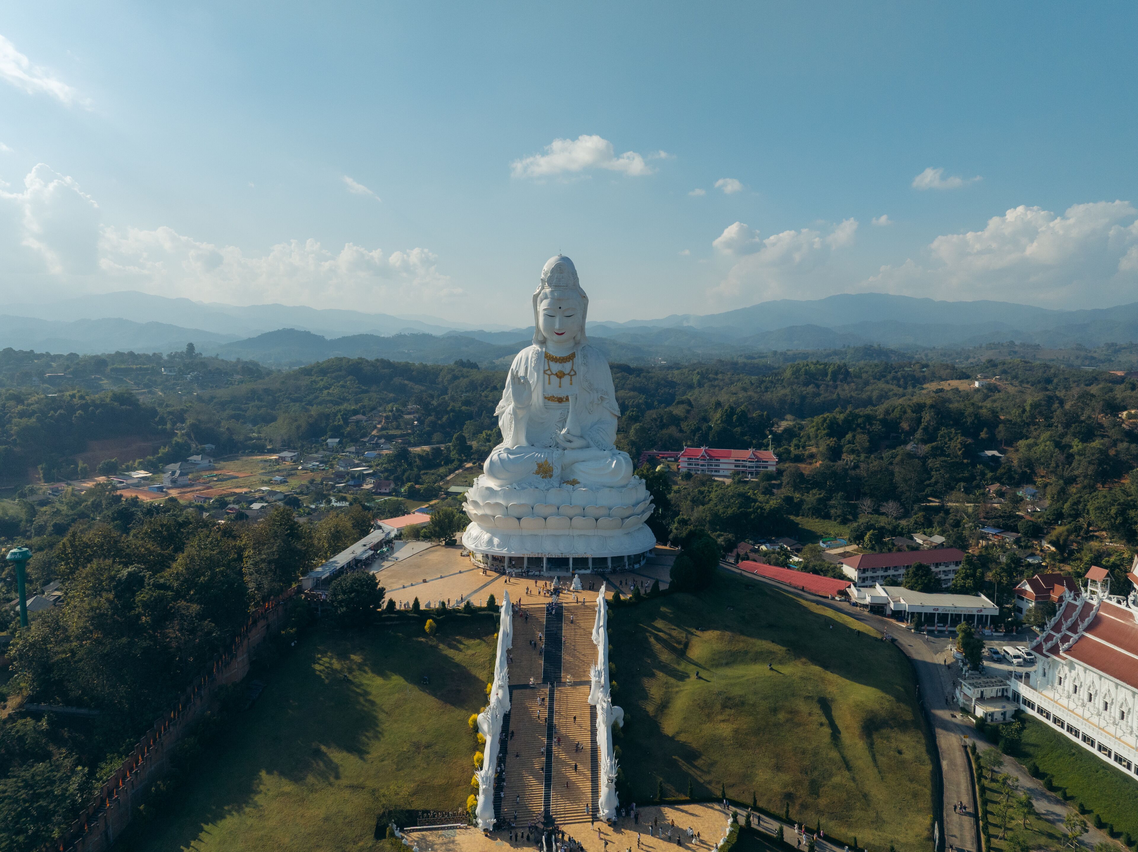 Aerial view of the majestic white Guan Yin statue standing tall against a backdrop of rolling green hills under a clear sky, Tambon Ban Du, Chang Wat Chiang Rai, Thailand.
