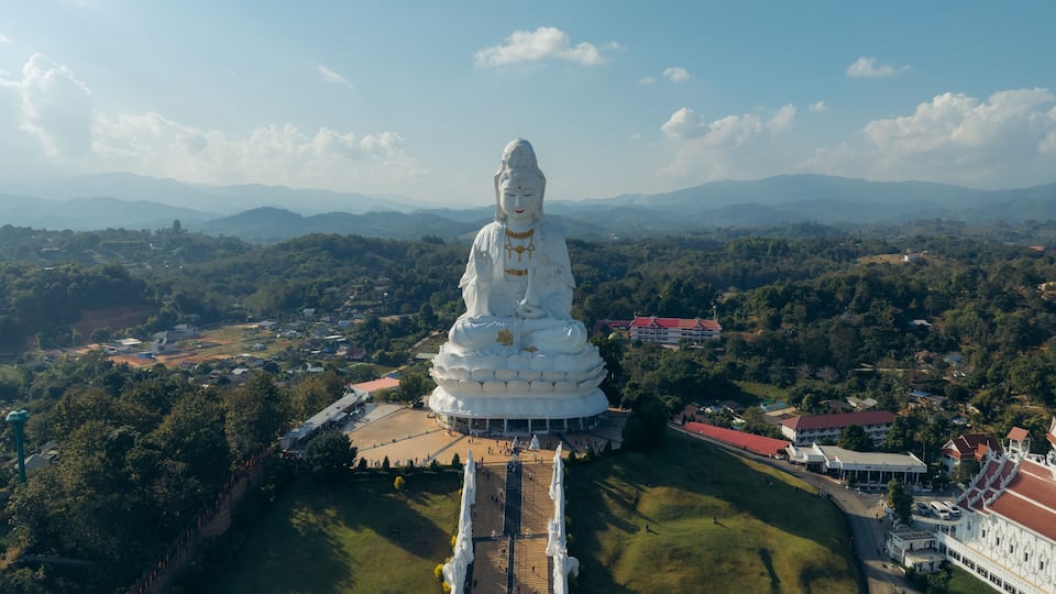 A large white Buddha statue surrounded by a well-maintained garden, with a view of distant mountains and a few buildings in the background.