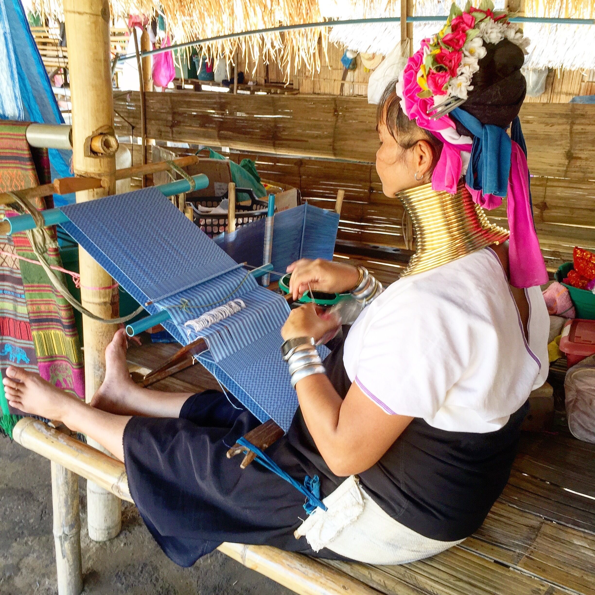 A person weaving on a loom in a traditional setting with thatched roofs and wooden structures.