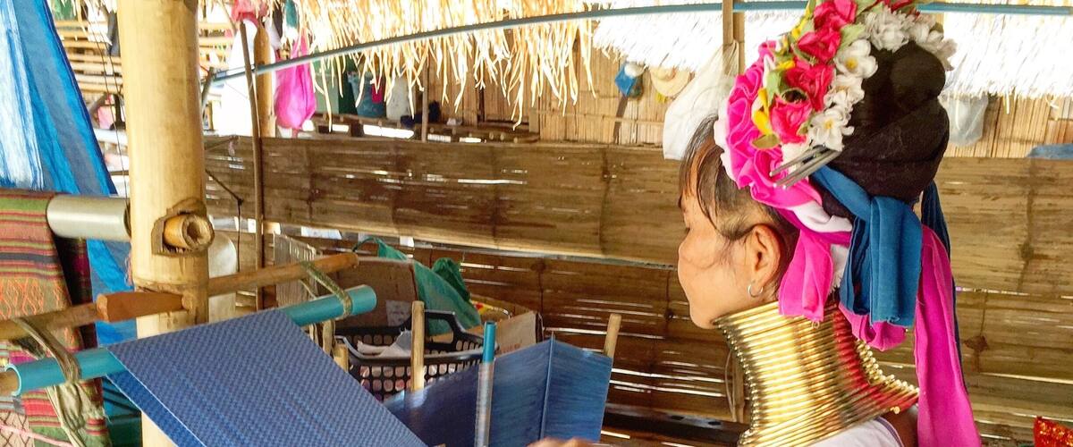 A person weaving on a loom in a traditional setting with thatched roofs and wooden structures.