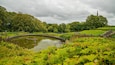 A pond surrounded by lush greenery, a stone staircase, and a distant church spire.