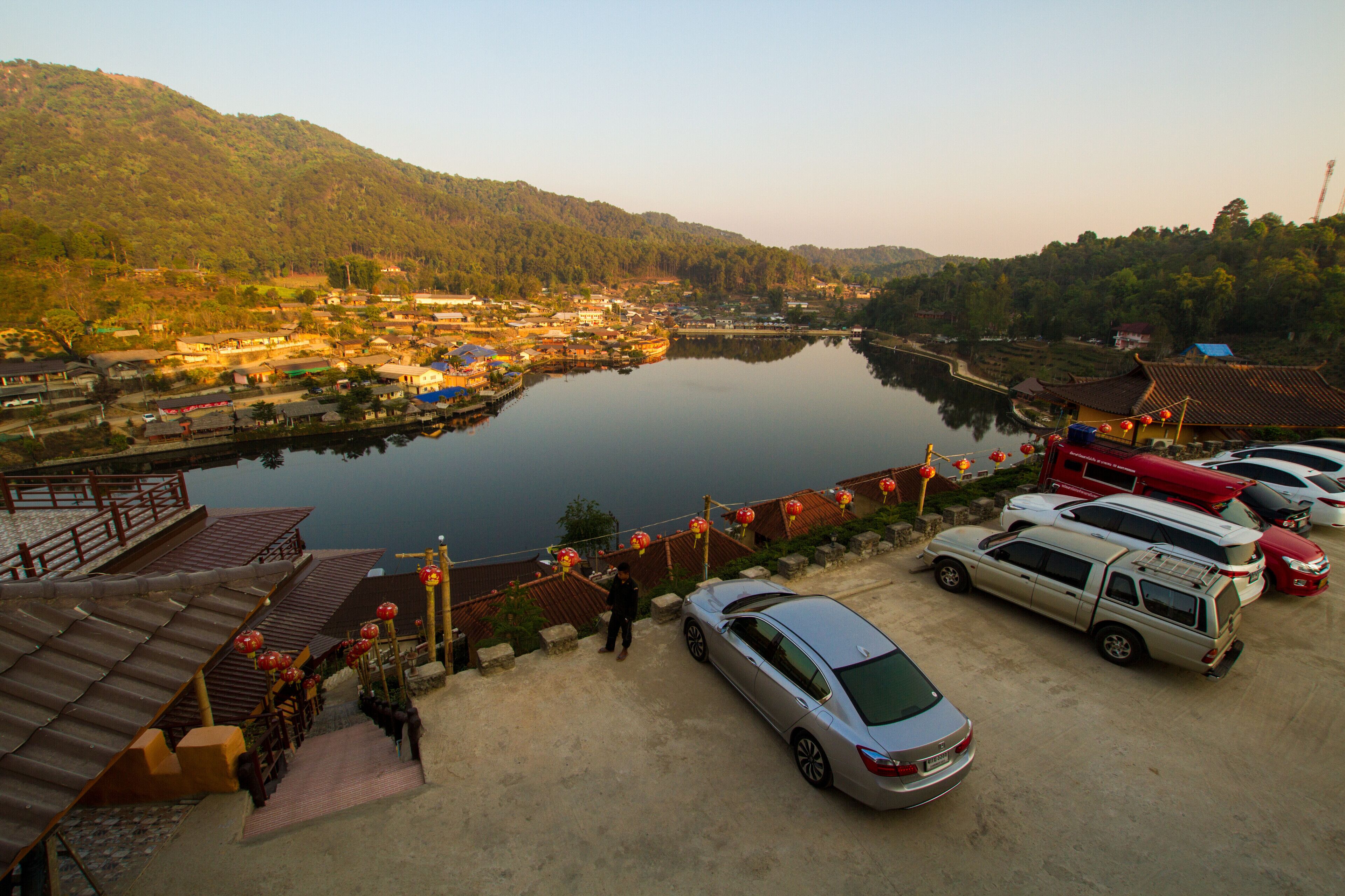 A serene lakeside village with traditional architecture, a parked car, and a red lantern-adorned pathway.