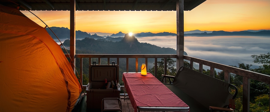 A tent, a table with a red checkered tablecloth, and a chair on a balcony overlooking a mountainous landscape at sunset.