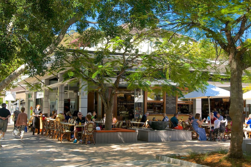 An outdoor café with tables and chairs under trees, surrounded by buildings.