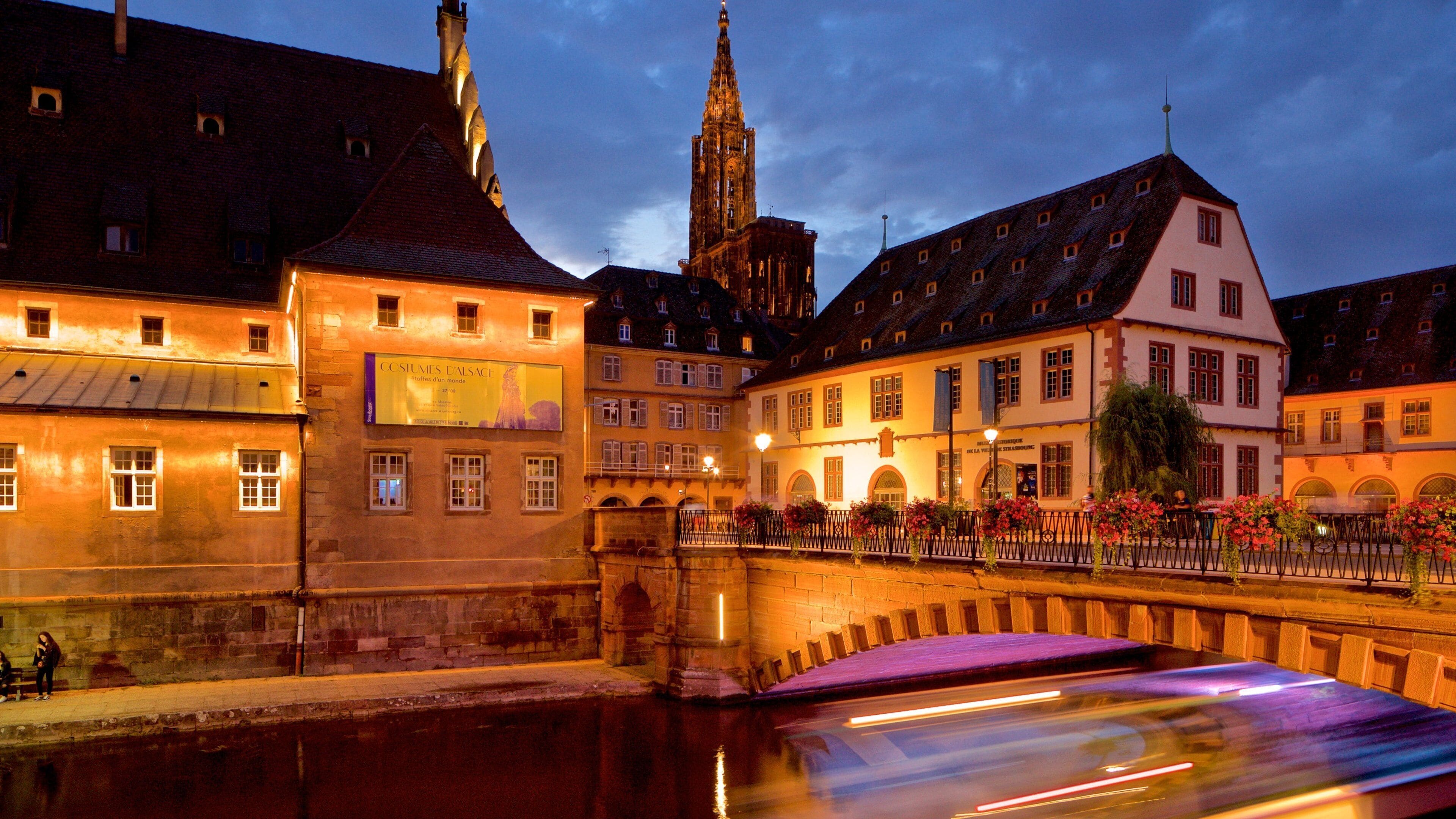 A historic European town with illuminated buildings, a prominent church tower, and a bridge over a river.