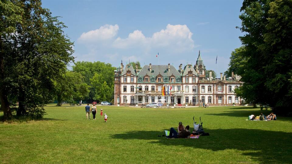 A large, historic building with a green roof and multiple flags, surrounded by a grassy park with people walking and sitting.