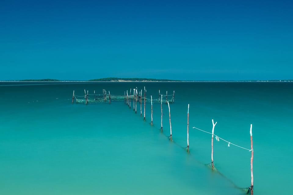 Un canal largo y estrecho con postes y cuerdas de madera, rodeado de aguas turquesas tranquilas y un cielo azul despejado.