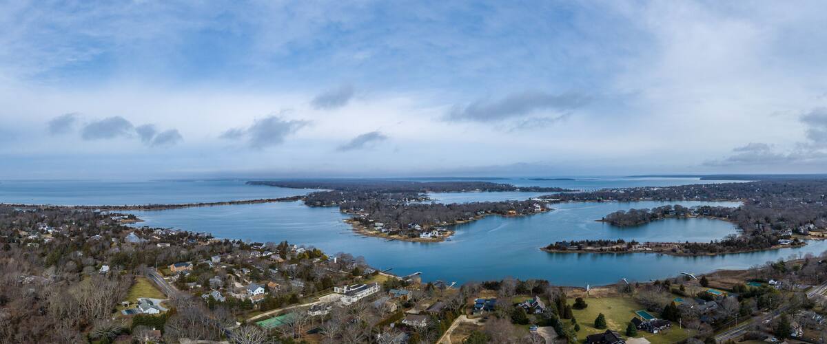 Aerial view of Sag Harbor Cove surrounded by residential properties, bare trees, and calm waters under a cloudy sky in Sag Harbor, NY, United States.