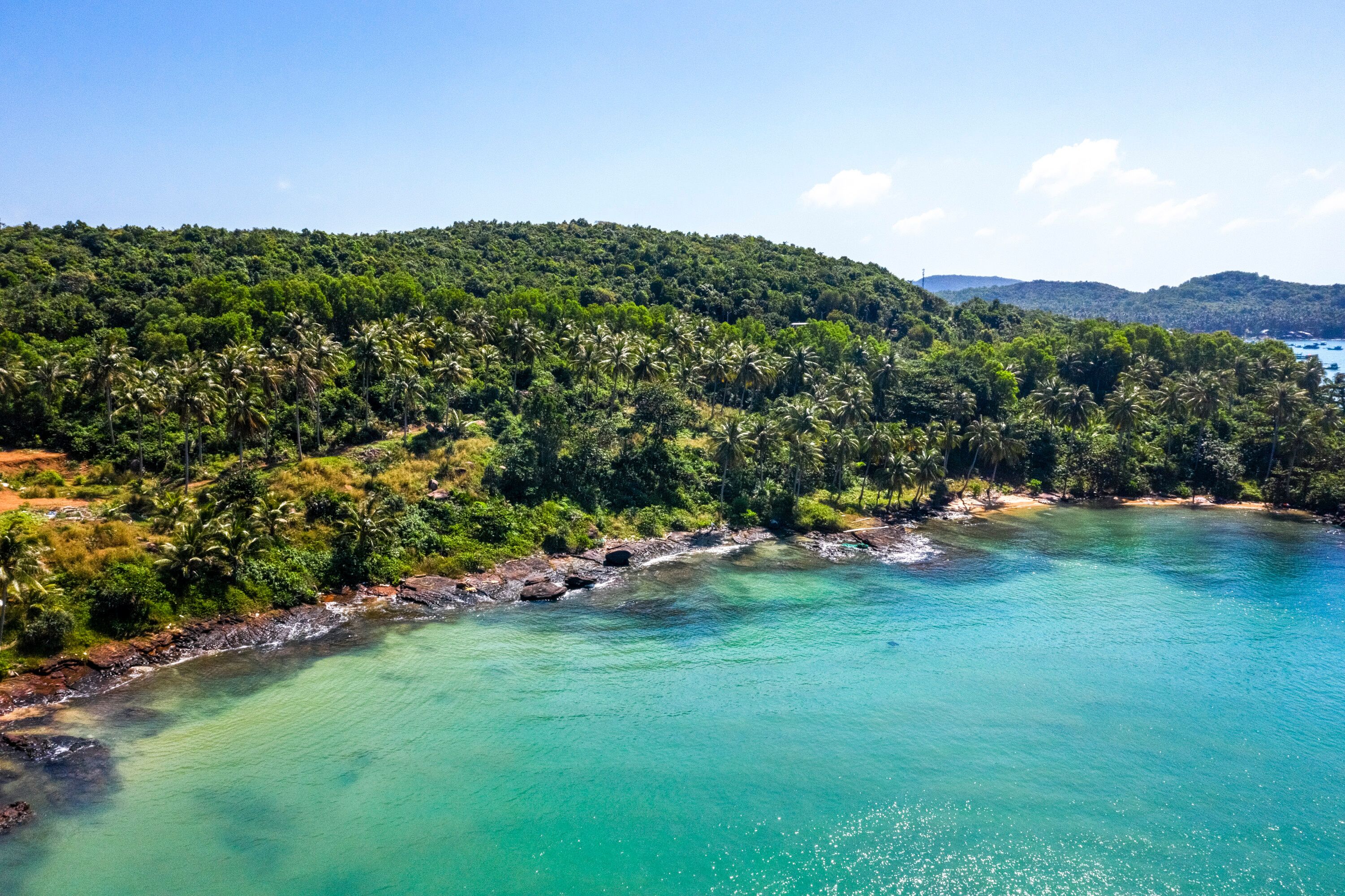 A coastal landscape with clear turquoise waters, a rocky shoreline, and dense green hills in the background.