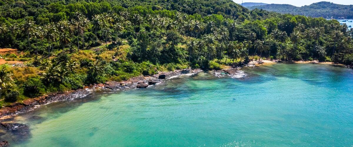 A coastal landscape with clear turquoise waters, a rocky shoreline, and dense green hills in the background.
