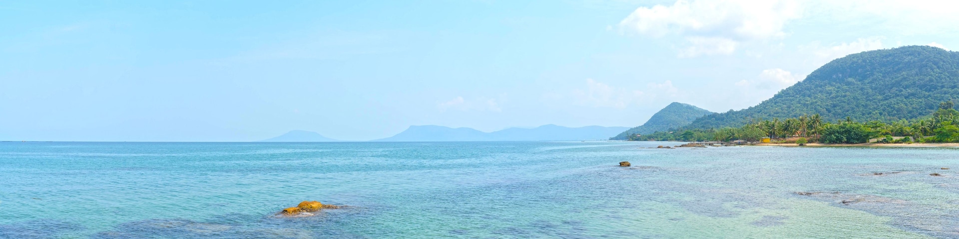 Una playa tropical con aguas turquesas cristalinas, afloramientos rocosos y colinas verdes exuberantes al fondo.