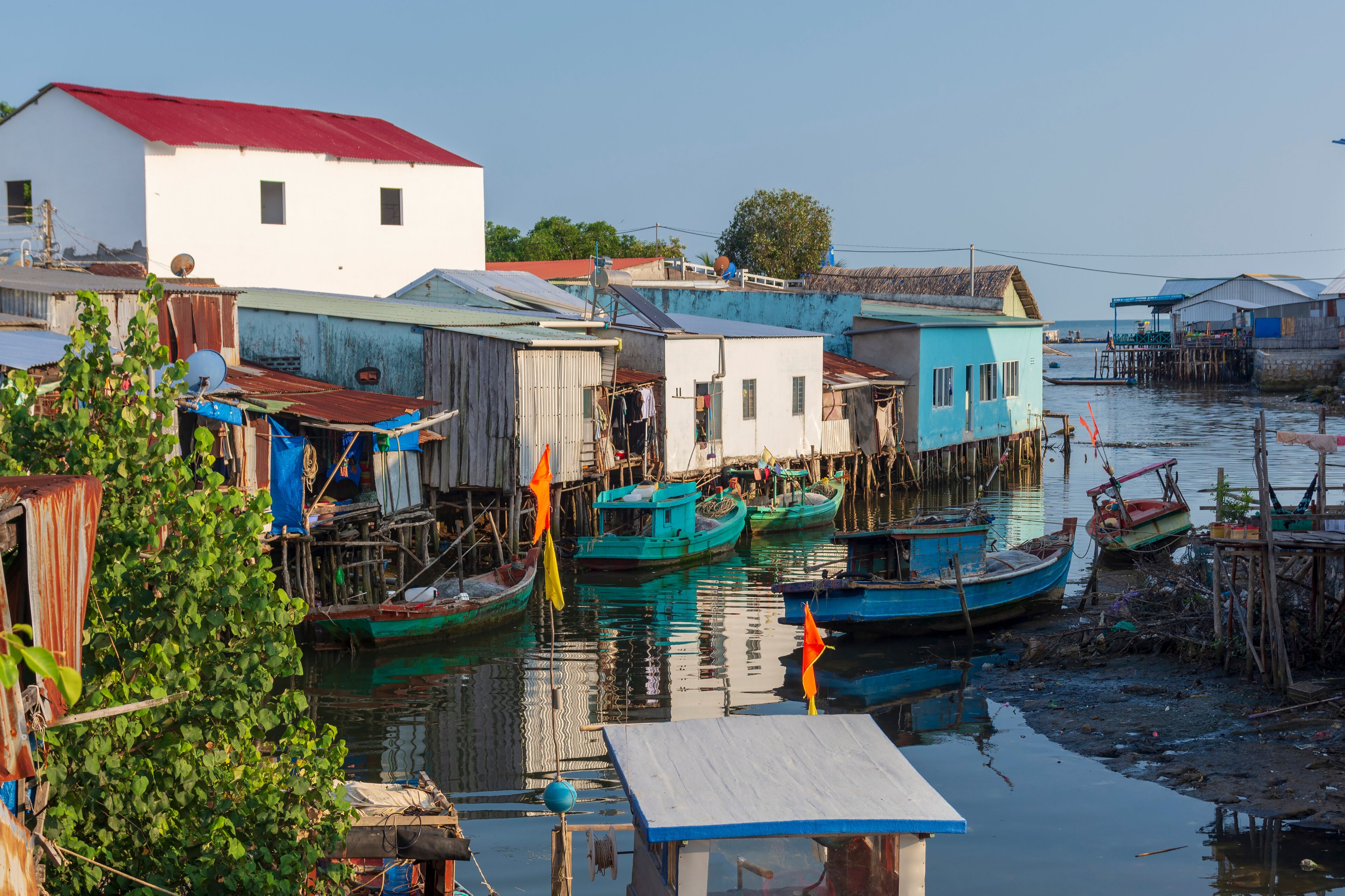A riverside settlement with boats docked at wooden piers, surrounded by small houses with corrugated metal roofs.
