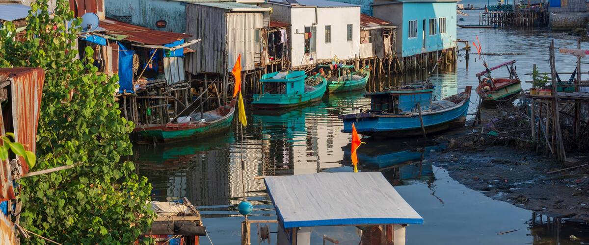 A riverside settlement with boats docked at wooden piers, surrounded by small houses with corrugated metal roofs.