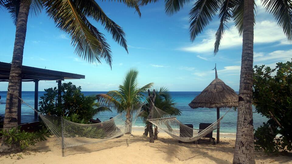 A beach with palm trees, a hammock, a thatched hut, and a wooden structure.