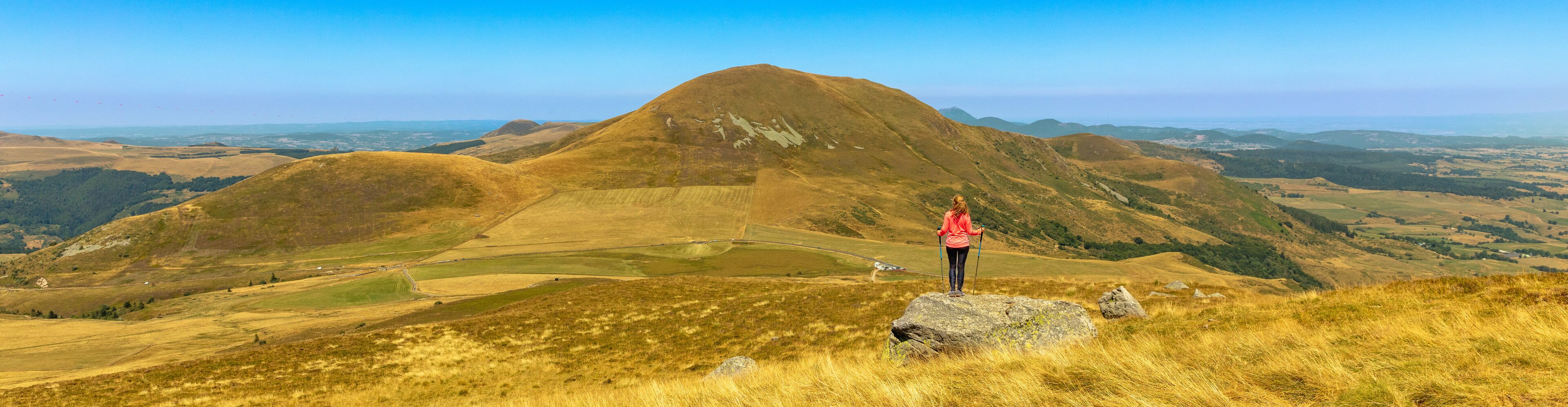 A hiker on a rocky outcrop overlooking a valley with rolling hills and distant mountains.
