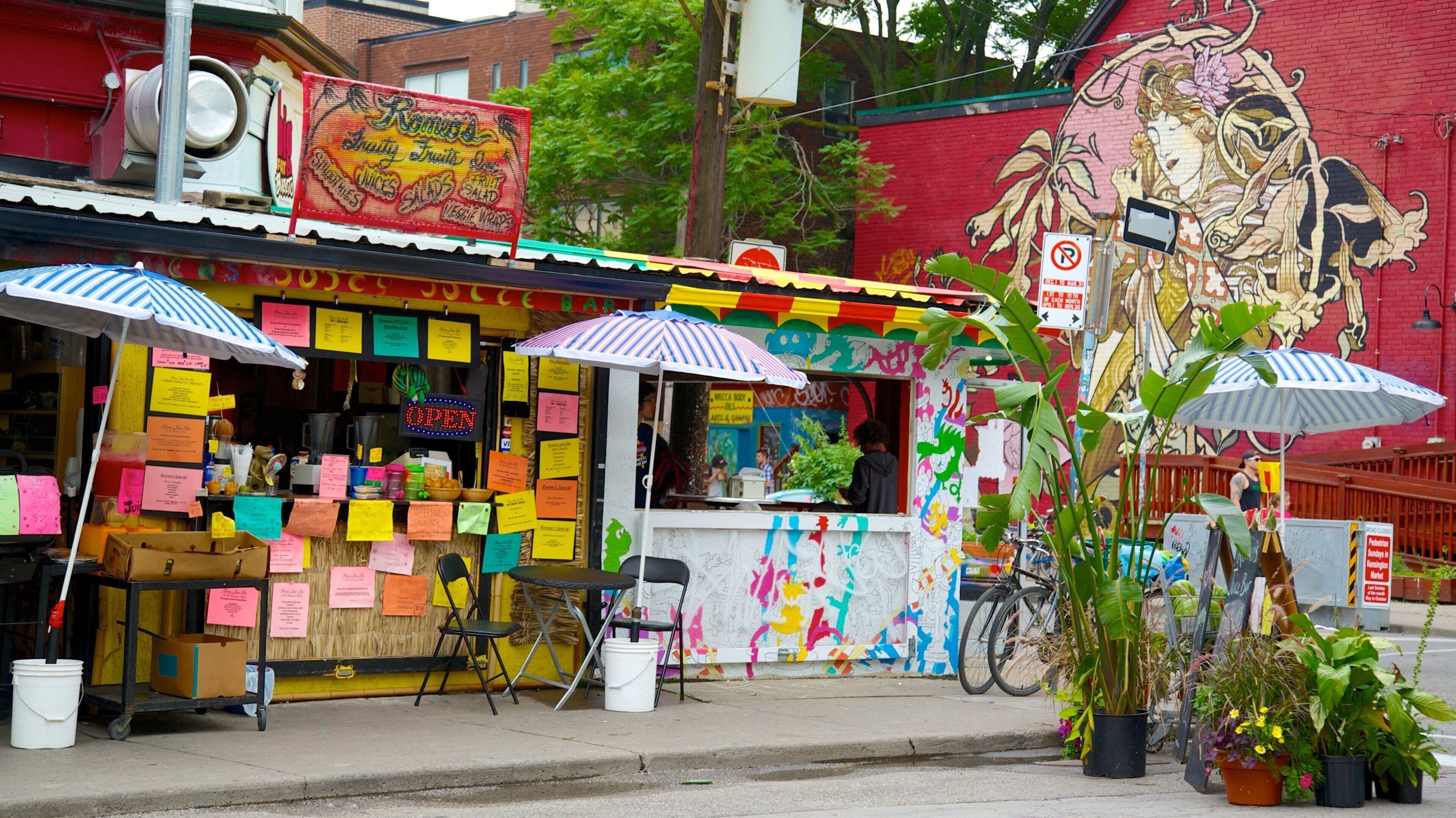 A colorful food cart with various items and notes, a mural on the wall, and a bicycle parked nearby.