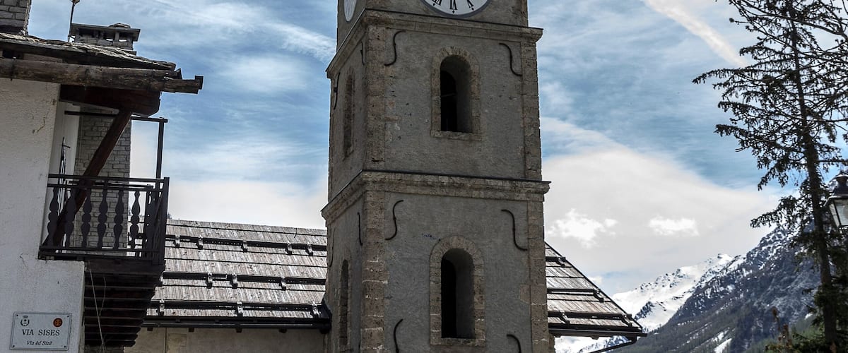 A clock tower with a weather vane, surrounded by buildings and a mountainous landscape.
