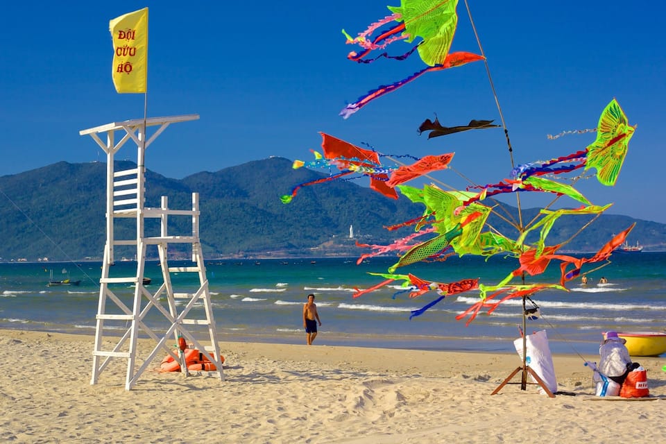 A beach with colorful kites, a lifeguard tower, and mountains in the background.