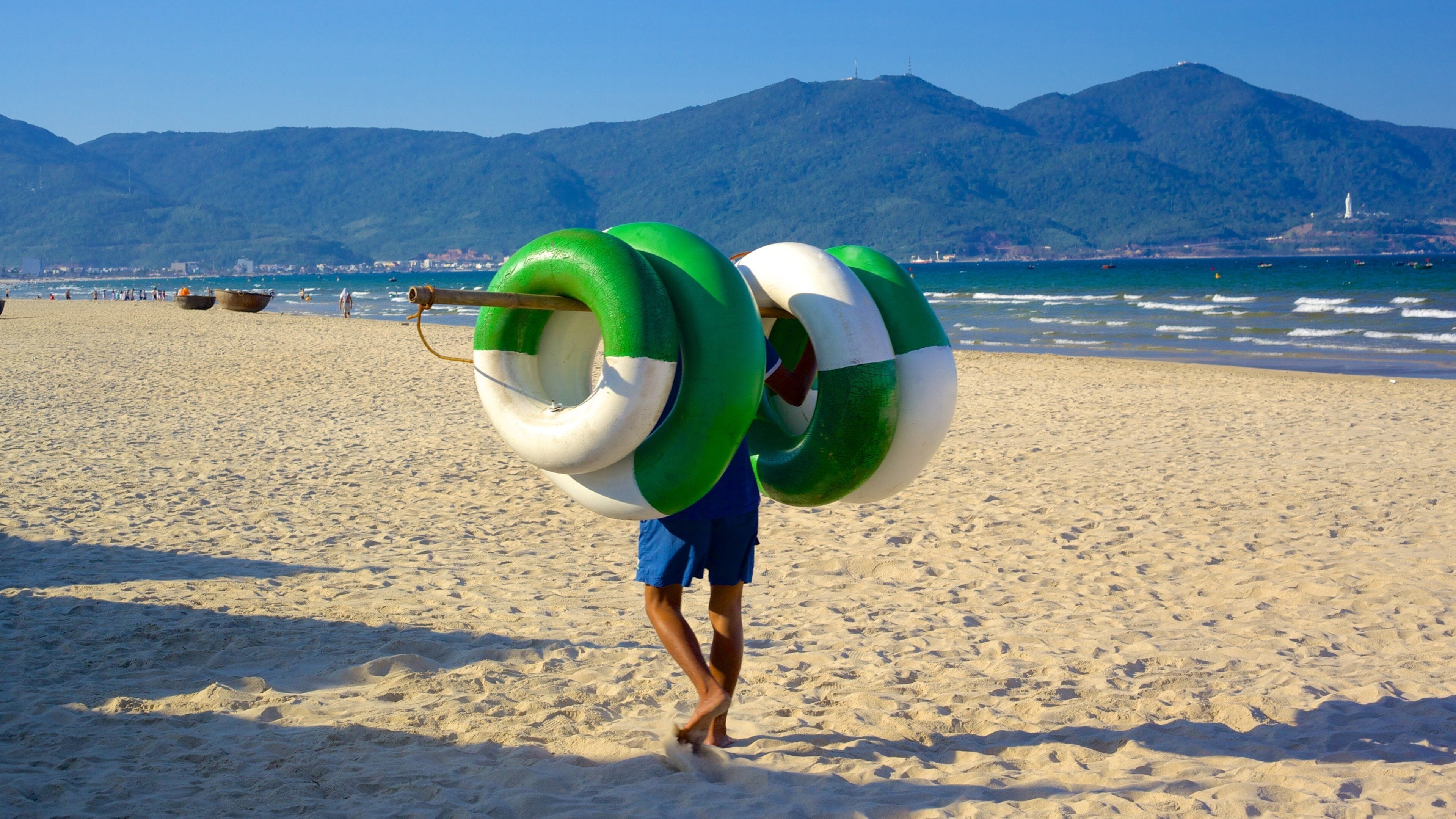 A person carrying large green and white inflatable rings on a sandy beach with mountains in the background.