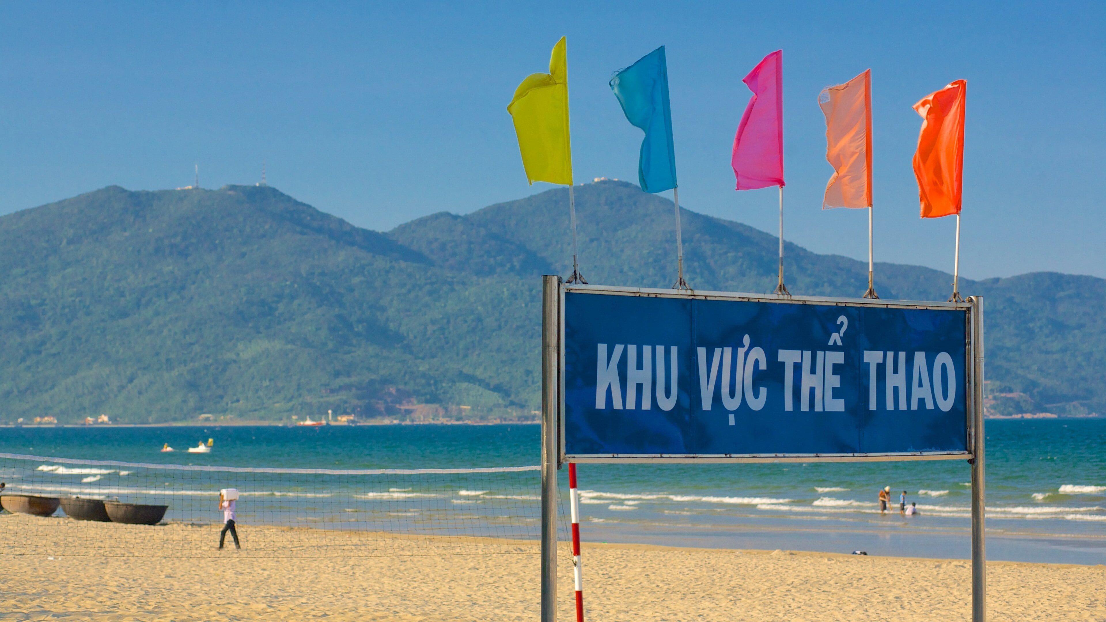 A beach with a sign reading "KHU VU C THE THAO" and colorful flags.
