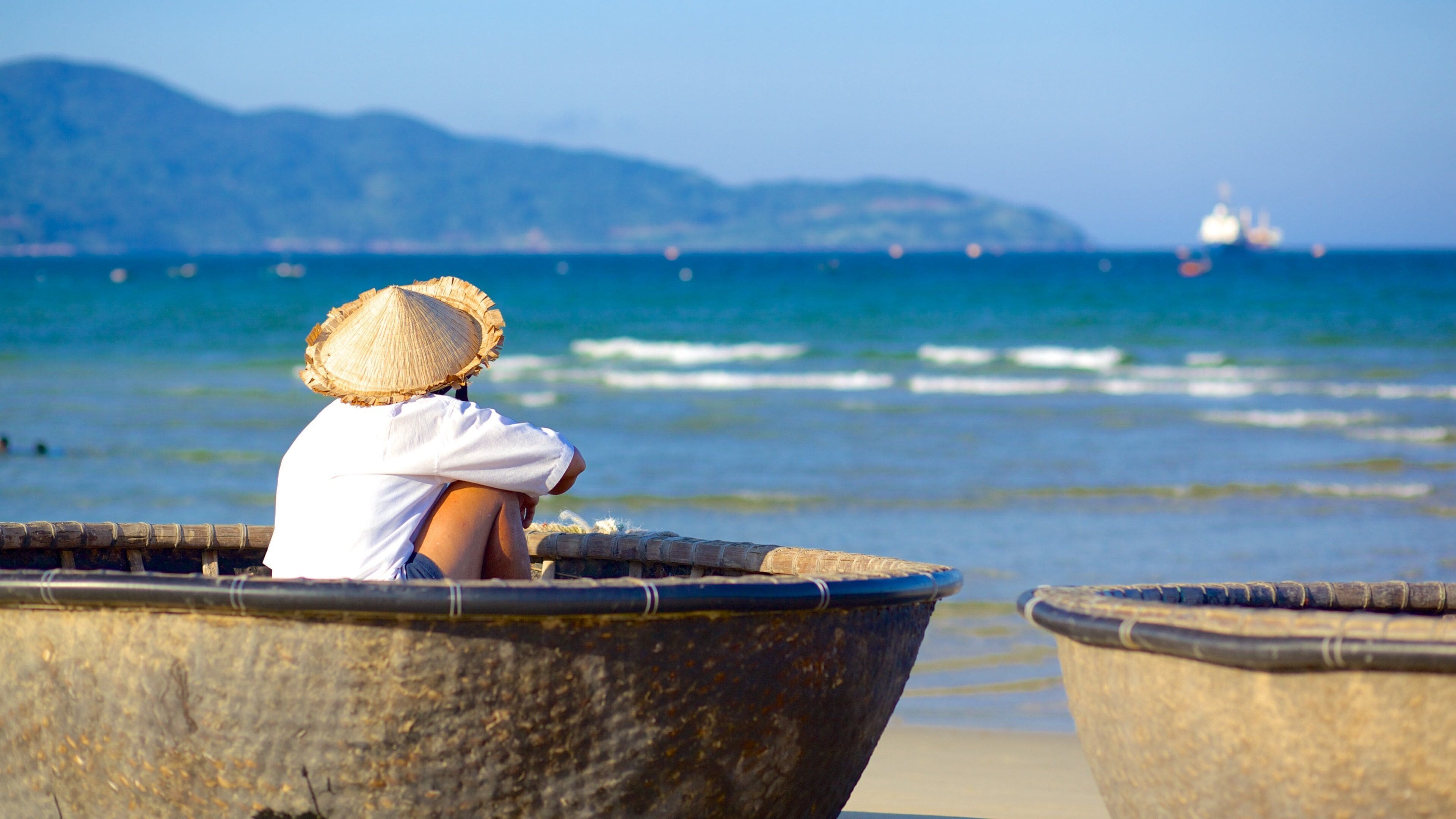 A person sitting in a traditional boat on a beach, with a calm sea and mountains in the background.