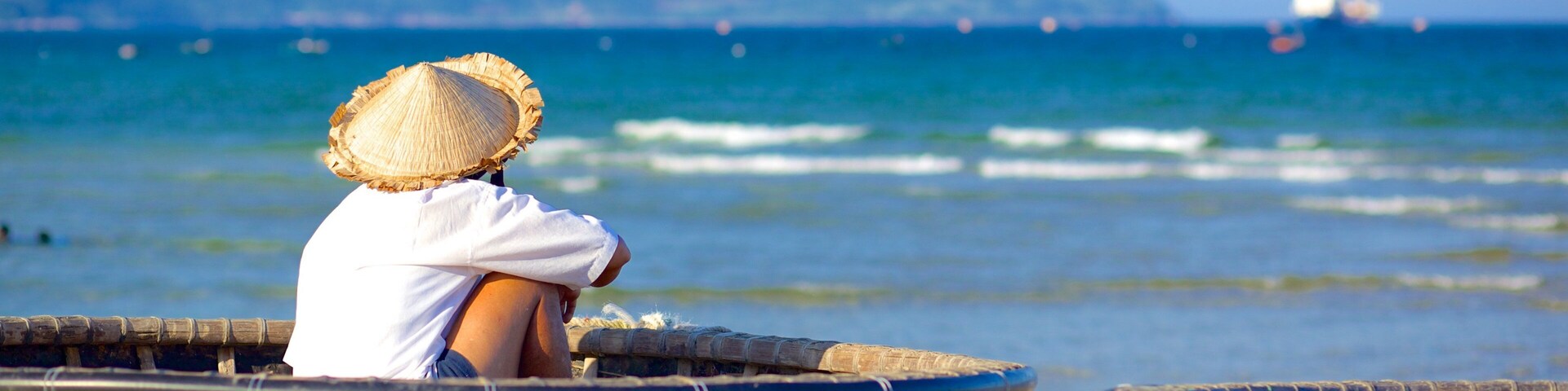 Una persona sentada en un bote tradicional en la playa, con un mar tranquilo y montañas al fondo.
