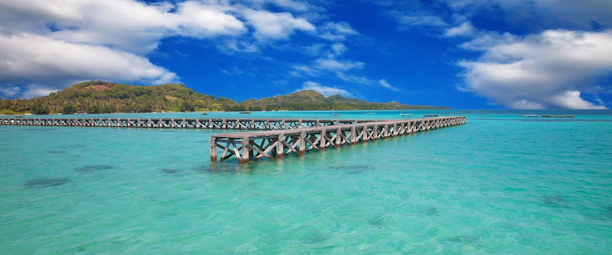 A wooden pier extending into turquoise waters, surrounded by a lush island and a partly cloudy sky.