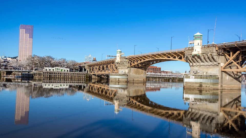 A bridge over a calm river with a clear reflection, a tall building, and a lighthouse.