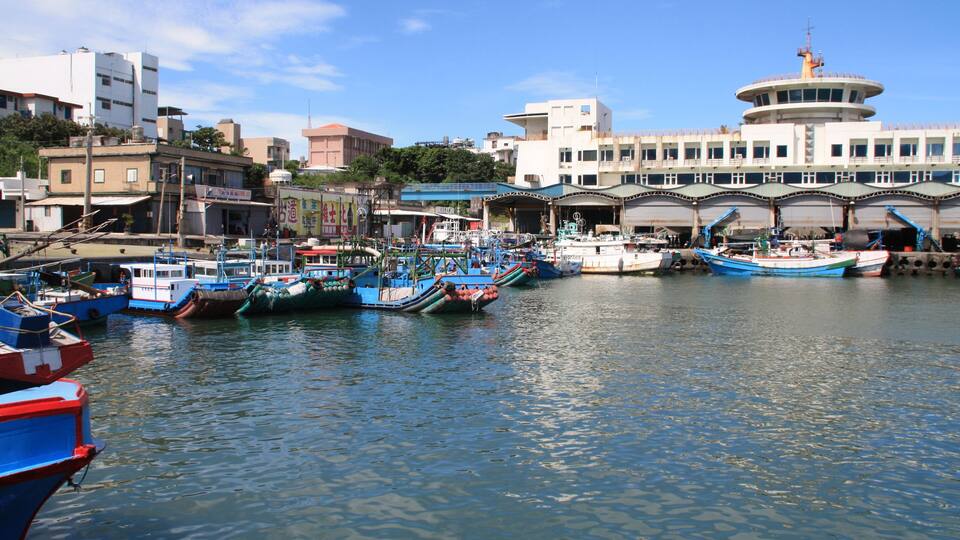 A harbor with multiple boats docked, a large building with a circular tower, and various other buildings in the background.